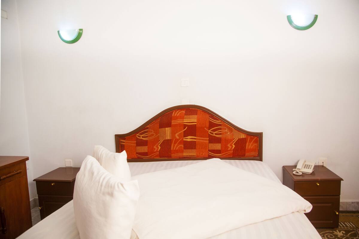 Una cama bien hecha con un cabecero de madera, dos mesitas de noche con teléfono y luces fijadas a la pared.