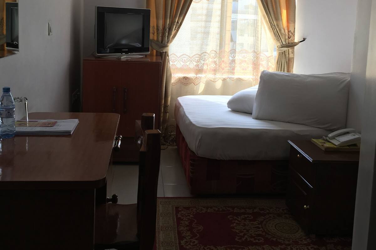 Habitación de hotel con cama, televisión sobre un mueble de madera, escritorio con silla y ventana con cortinas.