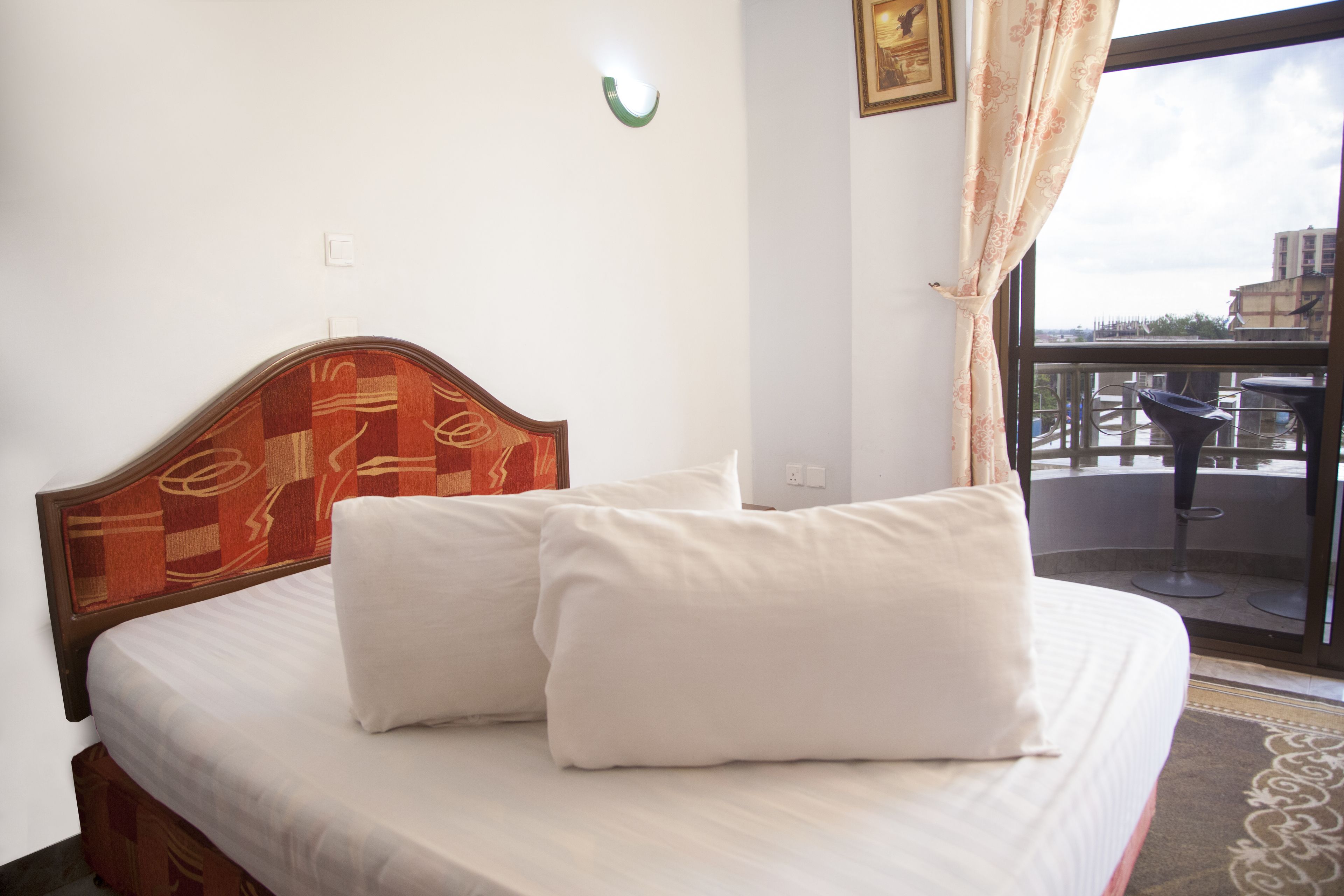 Habitación de hotel con cama, almohadas blancas, cabecera de madera, ventana con vistas al exterior y balcón pequeño con mesa y sillas.