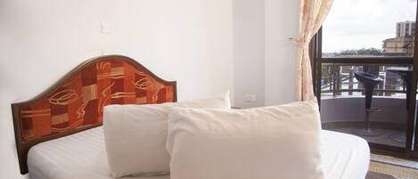 Habitación de hotel con cama, almohadas blancas, cabecera de madera, ventana con vistas al exterior y balcón pequeño con mesa y sillas.