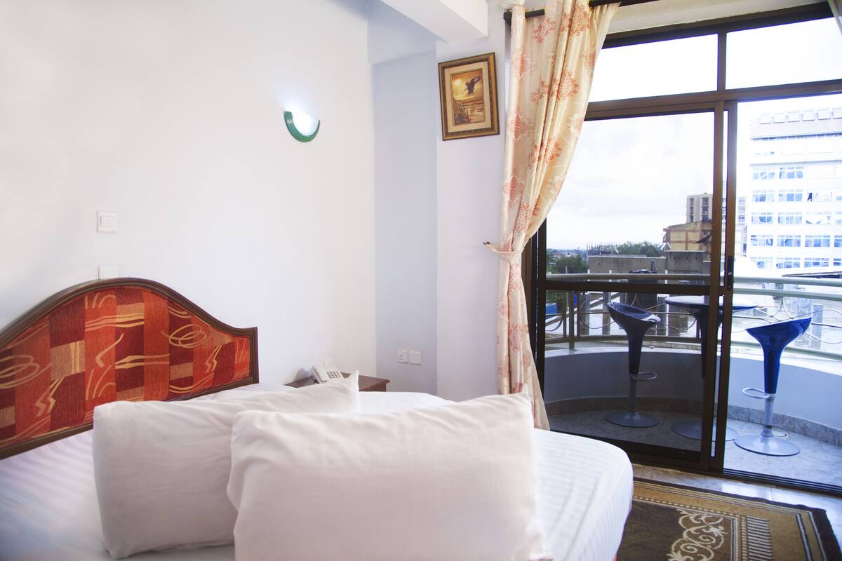 Habitación de hotel con cama, ventana con cortinas, vista de la ciudad y balcón con sillas.