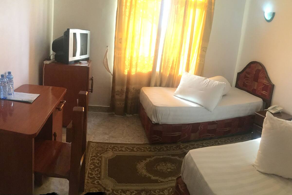 Habitación de hotel con escritorio de madera, televisor, dos camas con cabeceros rojos, alfombra estampada y ventana con cortinas amarillas.
