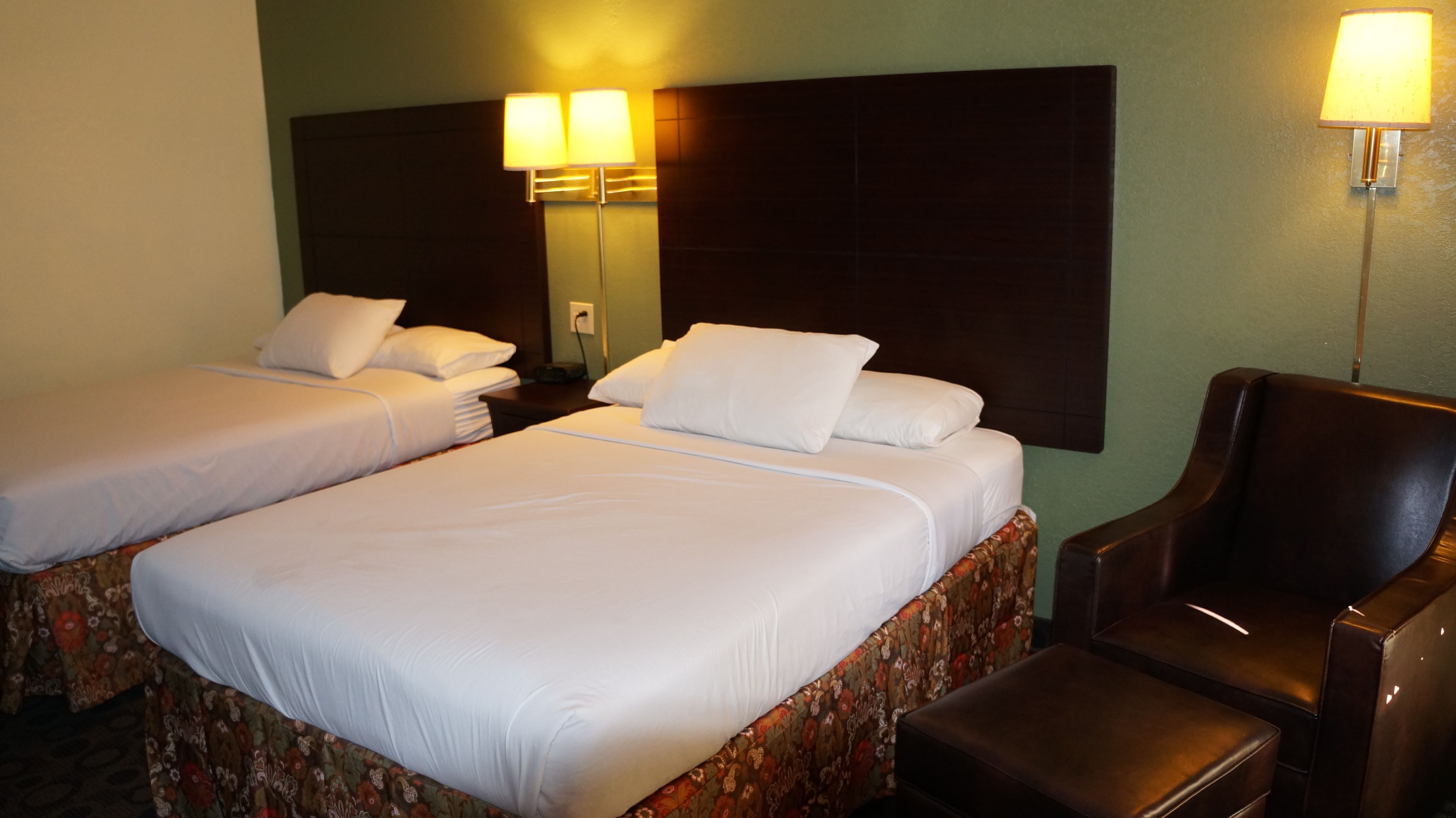 A hotel room with two single beds, a large headboard, a nightstand, a chair, and a lamp.