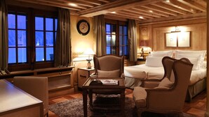 A hotel room with a bed, two armchairs, a small table, and a desk. There are windows with curtains, a wall clock, and a lamp on the nightstand. - Le Saint Joseph (Courchevel)