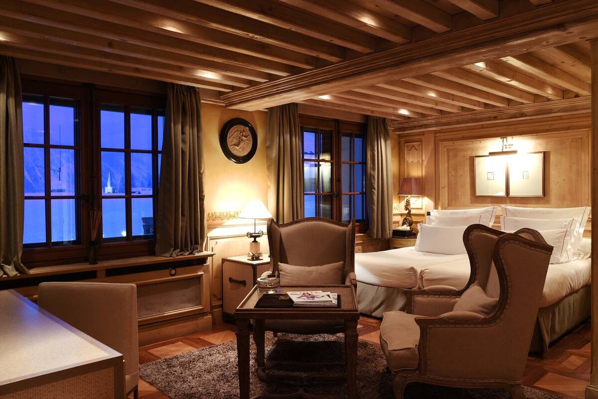 A hotel room with a bed, two armchairs, a small table, and a desk. There are windows with curtains, a wall clock, and a lamp on the nightstand.