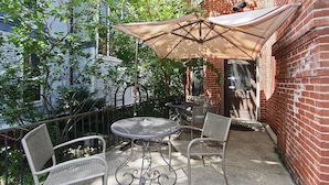 An outdoor patio with a round metal table and two chairs, a large beige umbrella, and a brick wall in the background.