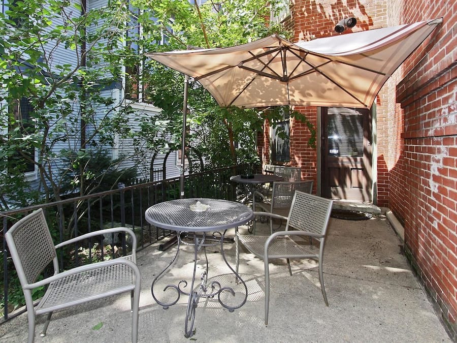 An outdoor patio with a round metal table and two chairs, a large beige umbrella, and a brick wall in the background.