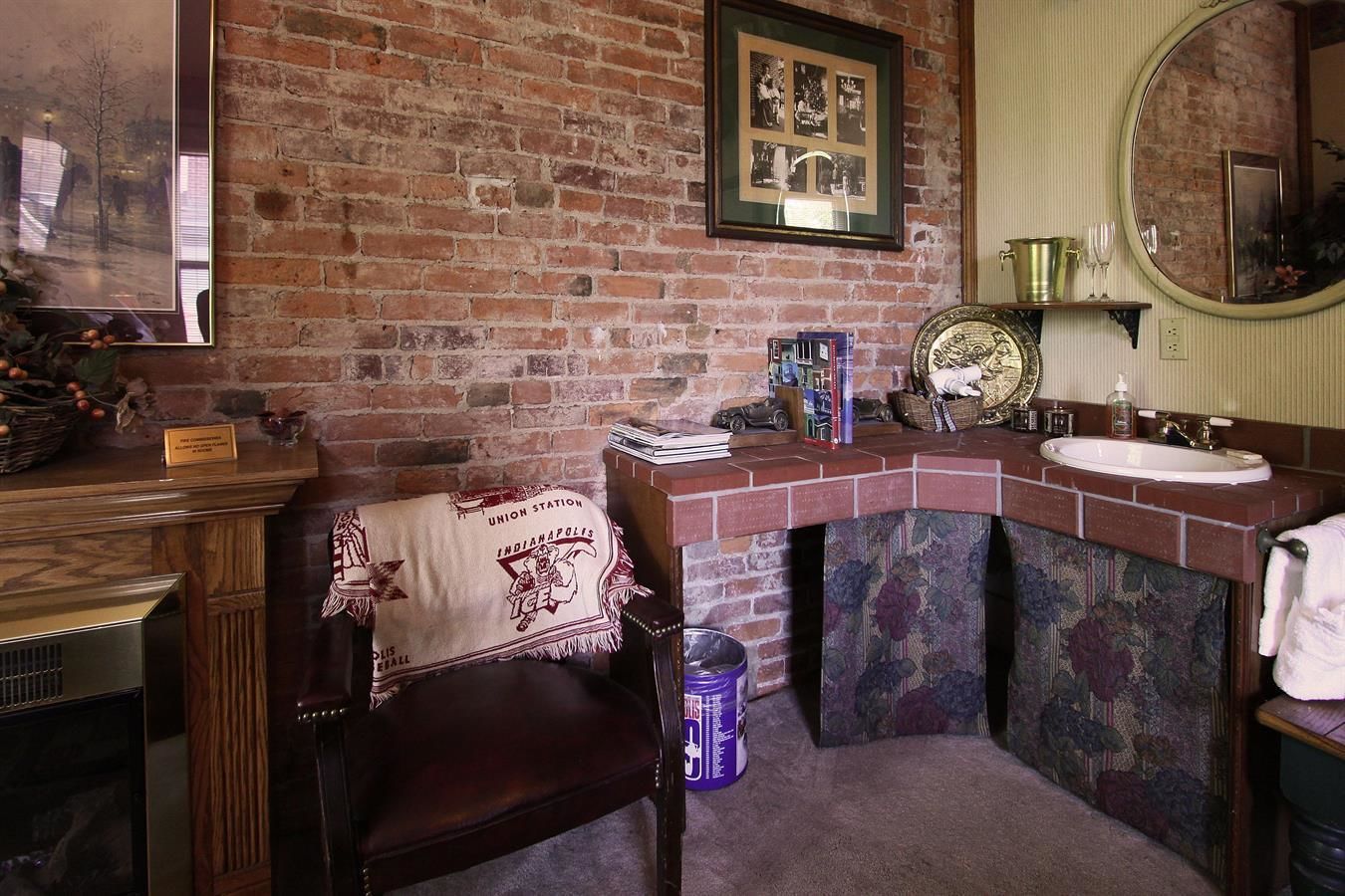 A bathroom with a brick wall, a wooden chair, a sink, a mirror, and a framed picture on the wall.