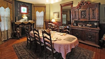Une salle à manger avec une table ronde dressée pour quatre personnes, entourée de chaises en bois. On y trouve une cheminée avec un manteau de cheminée, un grand miroir et un meuble contenant des objets décoratifs.
