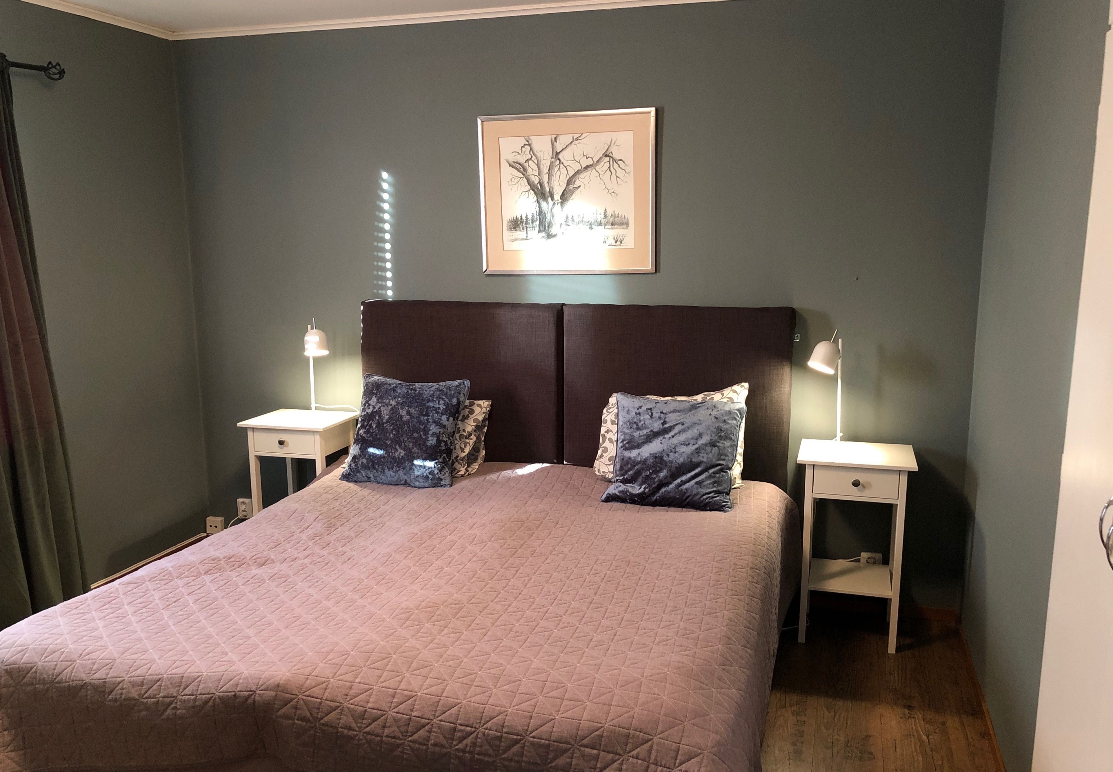 A neatly made bed with a quilted cover, two bedside tables with lamps, a framed picture on the wall, and a window with curtains.