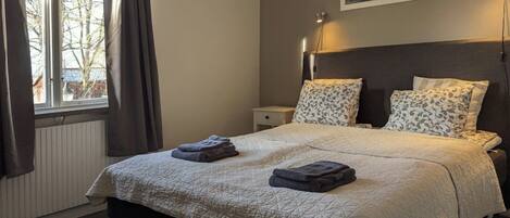 A bedroom with a bed, a nightstand, a lamp, a window with curtains, and a painting on the wall.