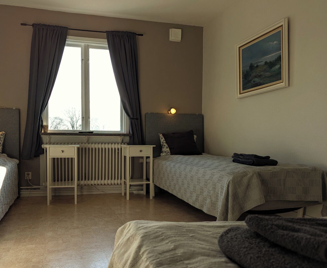 Un dormitorio con dos camas, una ventana con cortinas, un radiador, una mesita de noche y un cuadro en la pared.