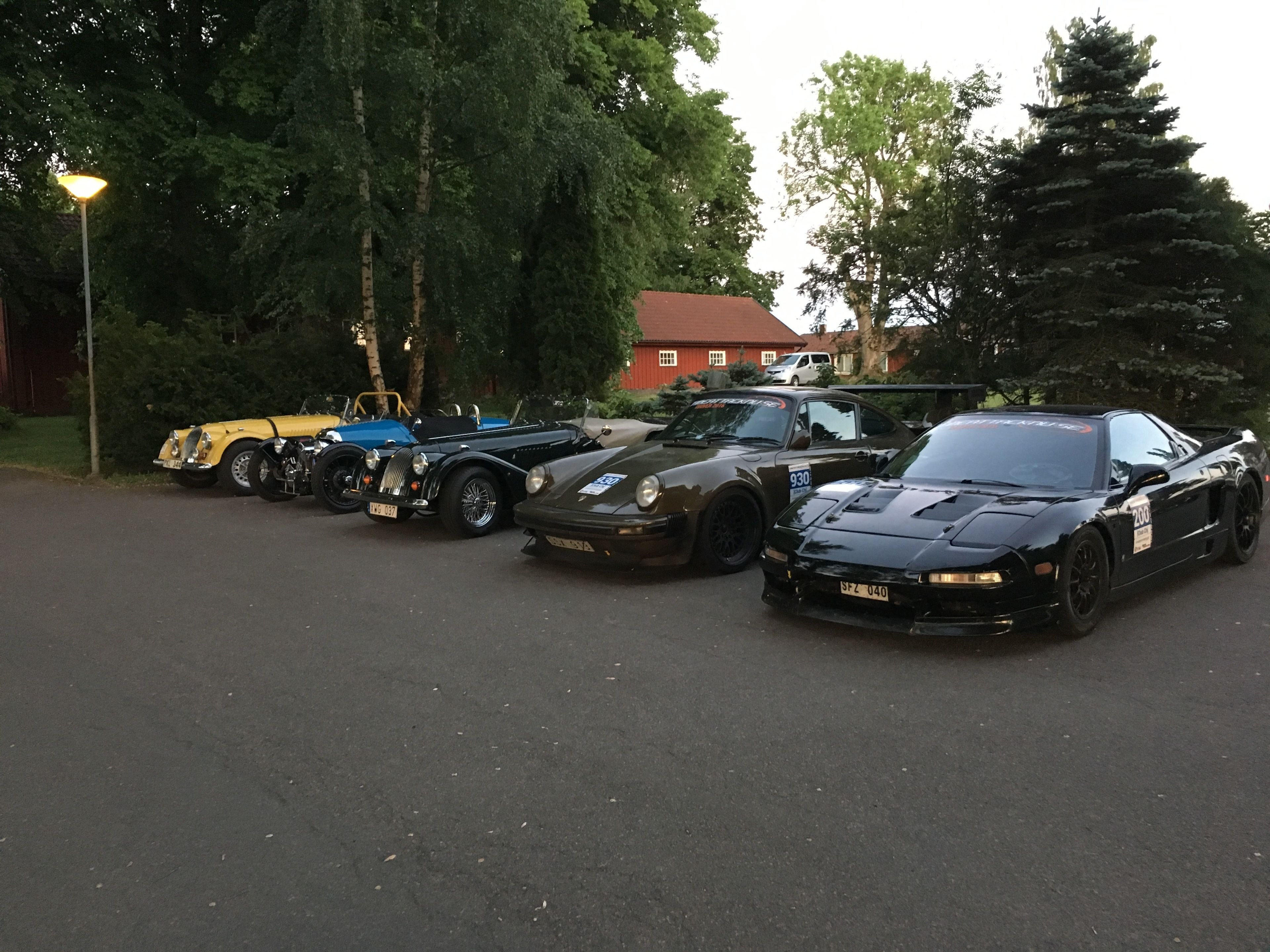 A row of vintage and modern sports cars parked in a lot with trees and a building in the background.