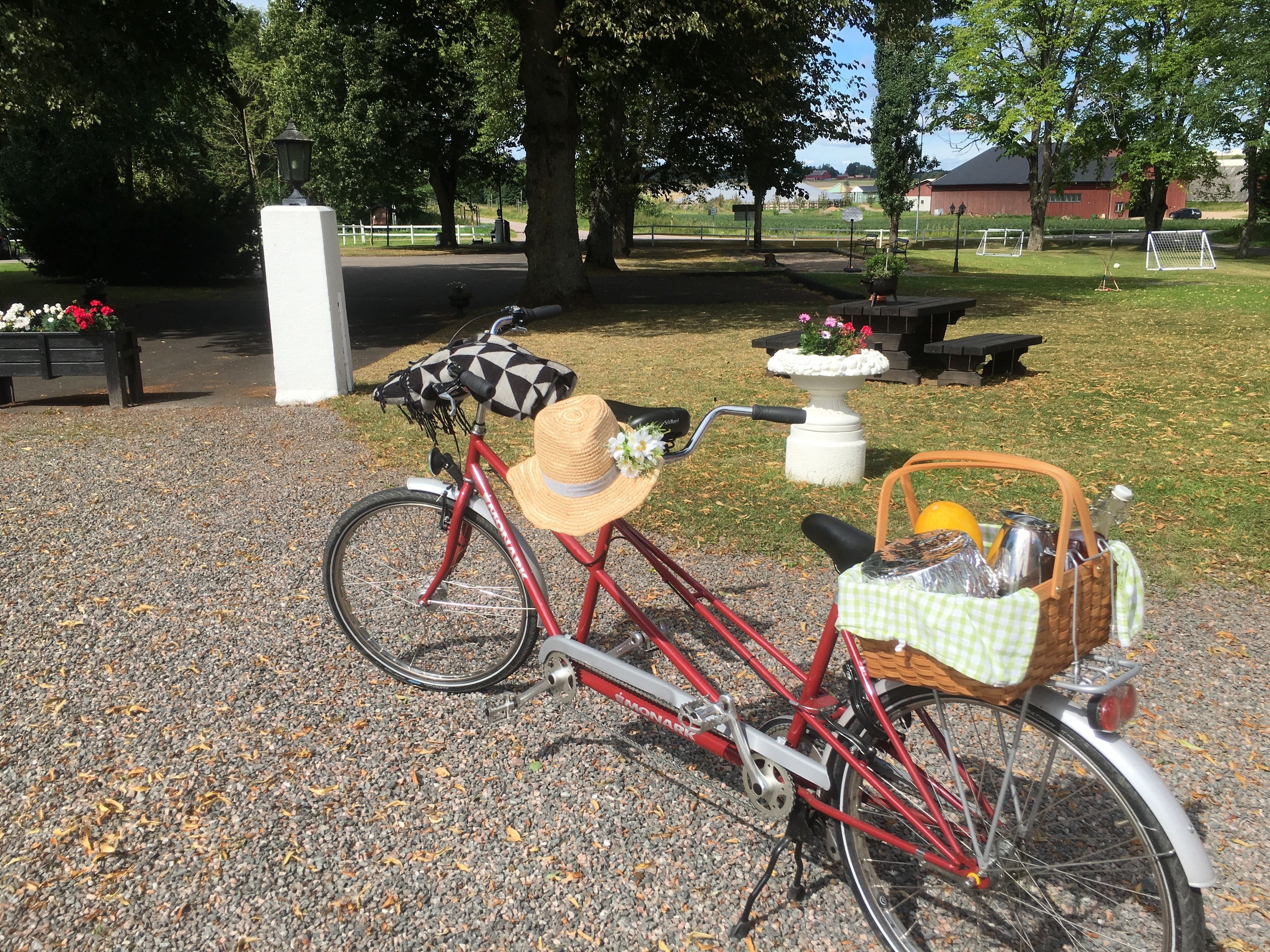A red bicycle with a basket and a hat on the seat, parked on a gravel path in a park.