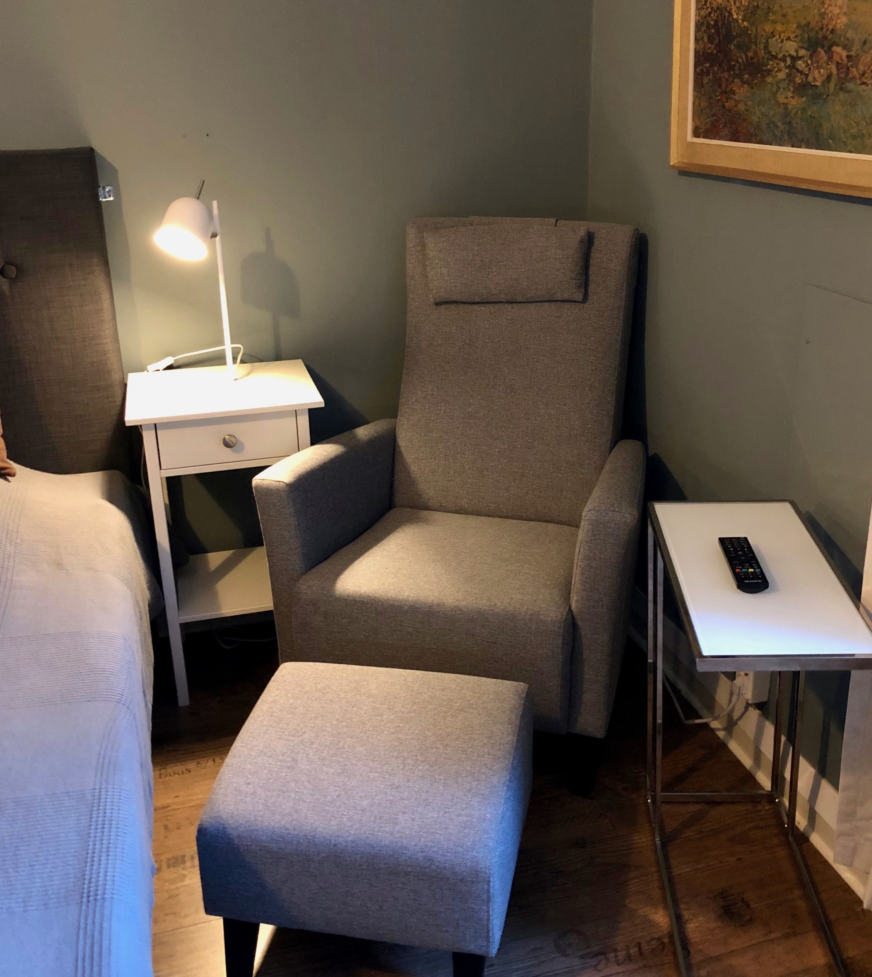 A room with a grey armchair, a small table with a lamp, a bedside table with a remote control, and a wooden floor.