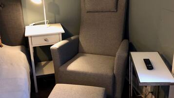 A room with a grey armchair, a small table with a lamp, a bedside table with a remote control, and a wooden floor.