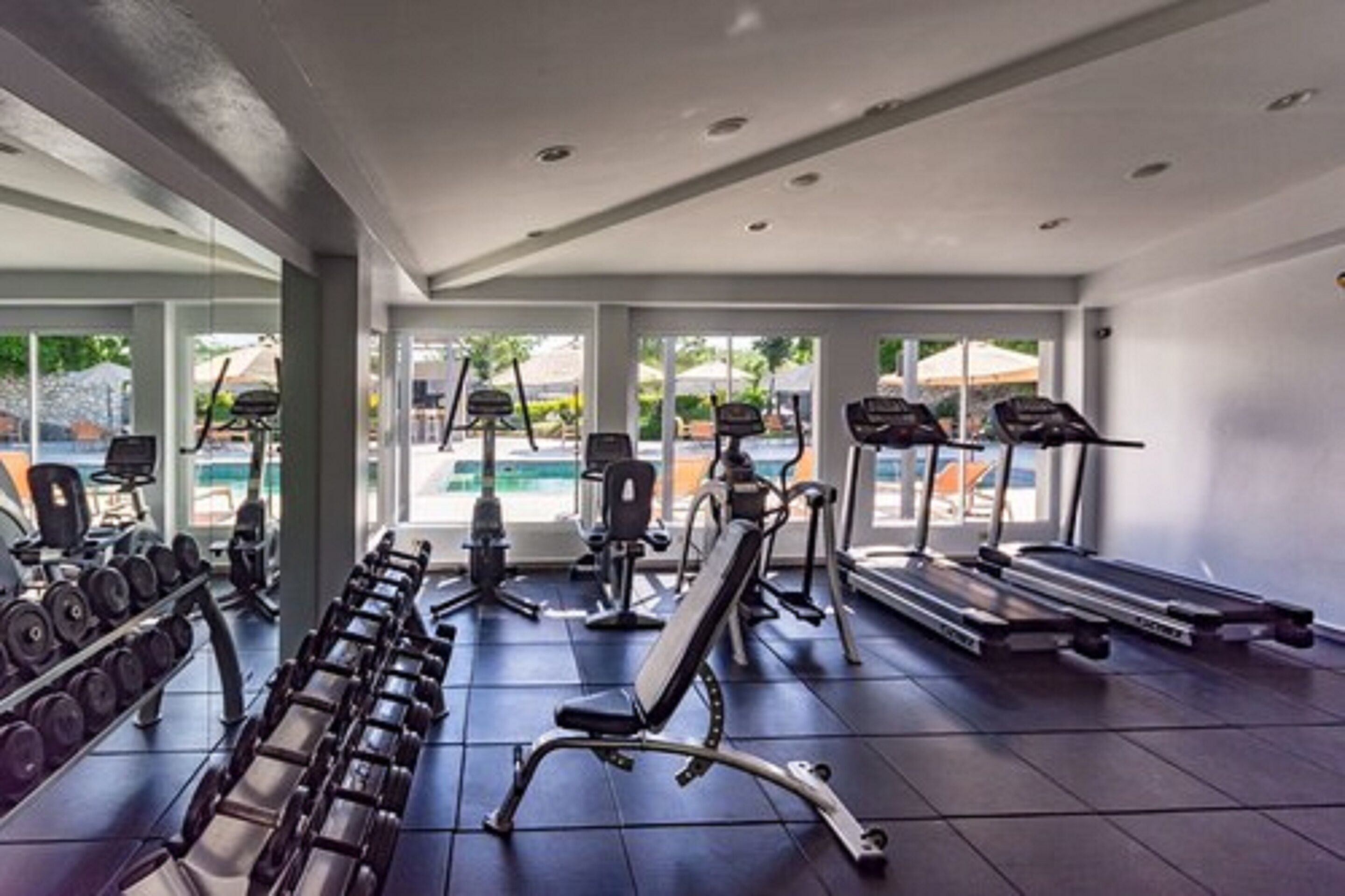 Ein Fitnessraum mit Trainingsgeräten, Laufband und Blick auf einen Pool.