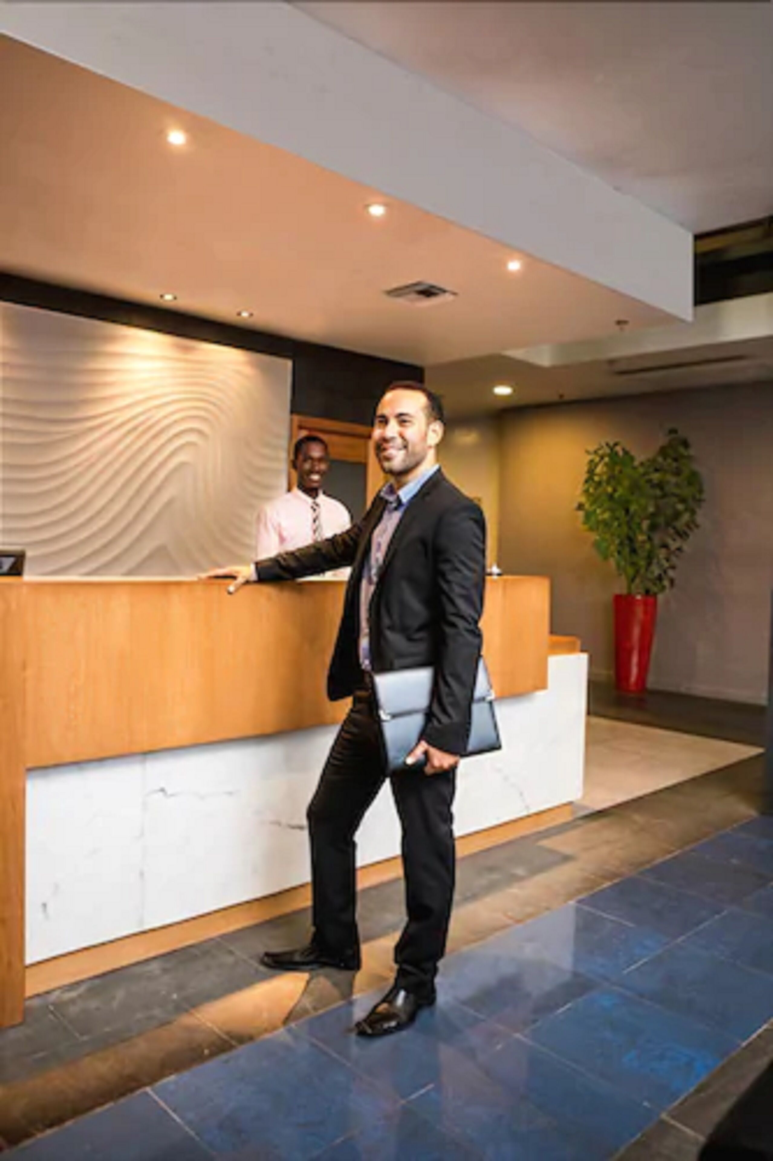 A modern reception area with a wooden counter, a large potted plant, and a person standing with a briefcase.