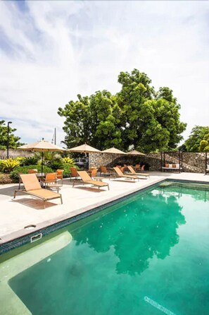 Una piscina con sillas de descanso y sombrillas, rodeada de follaje y un muro de piedra.