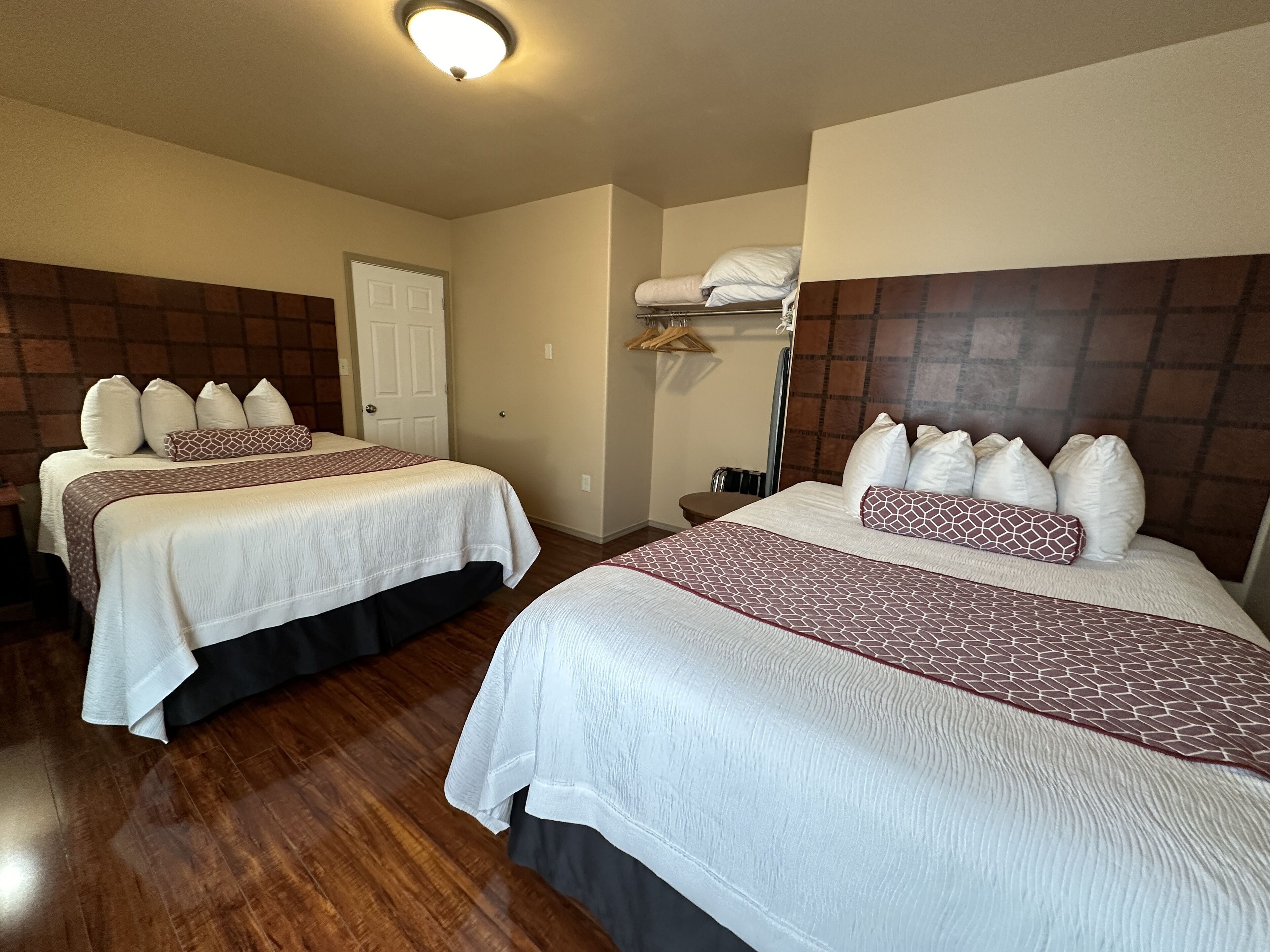 Dos camas dobles con sábanas blancas y almohadas estampadas, cabeceros de madera, una puerta blanca y una mesita de noche con una lámpara.