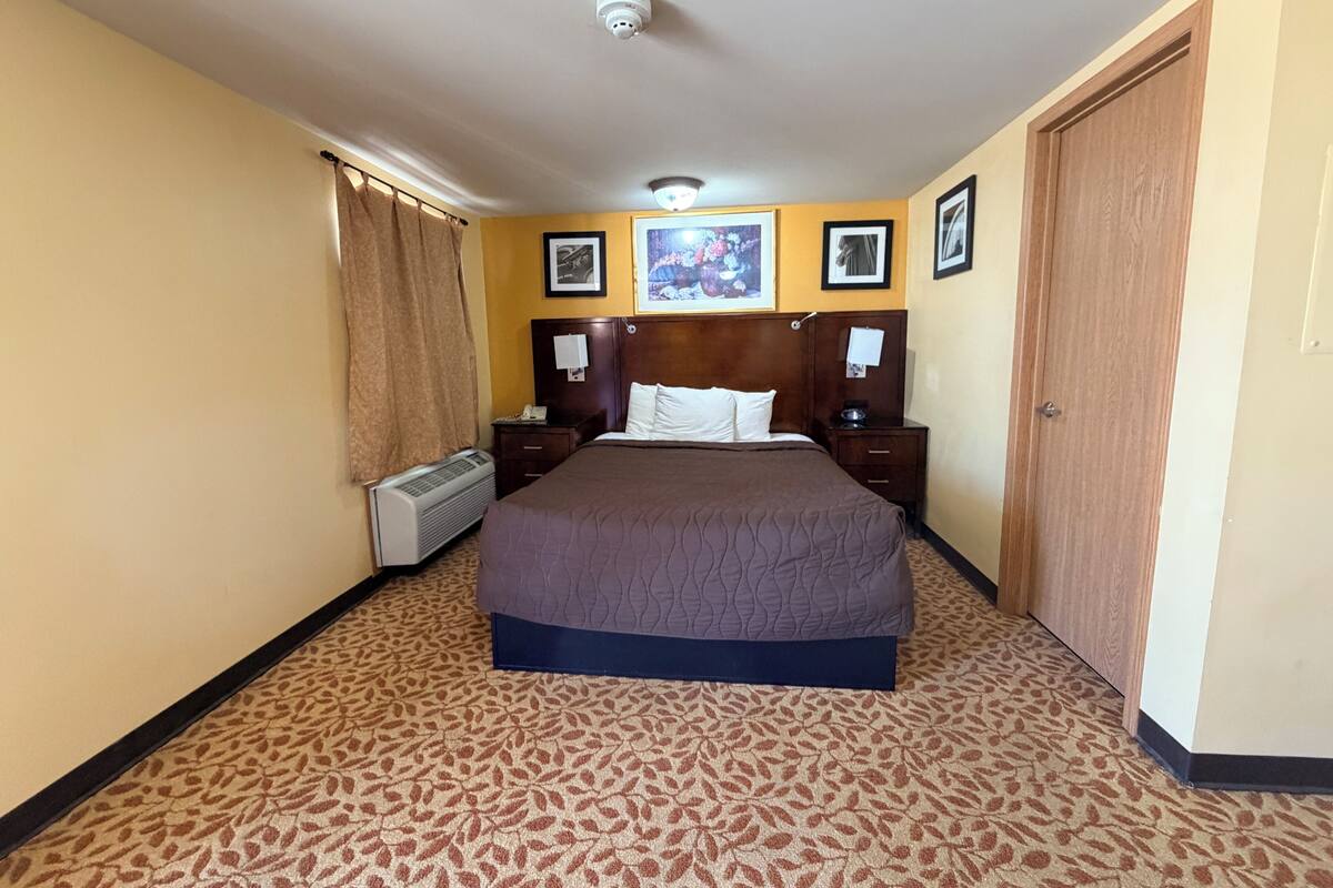 Habitación de hotel con cama, mesita de noche, calentador en la pared, puerta y luz de techo.