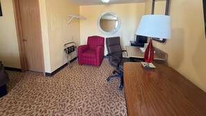 A hotel room with a wooden door, a red armchair, a black office chair, a desk with a lamp, a round mirror, and a ceiling light.