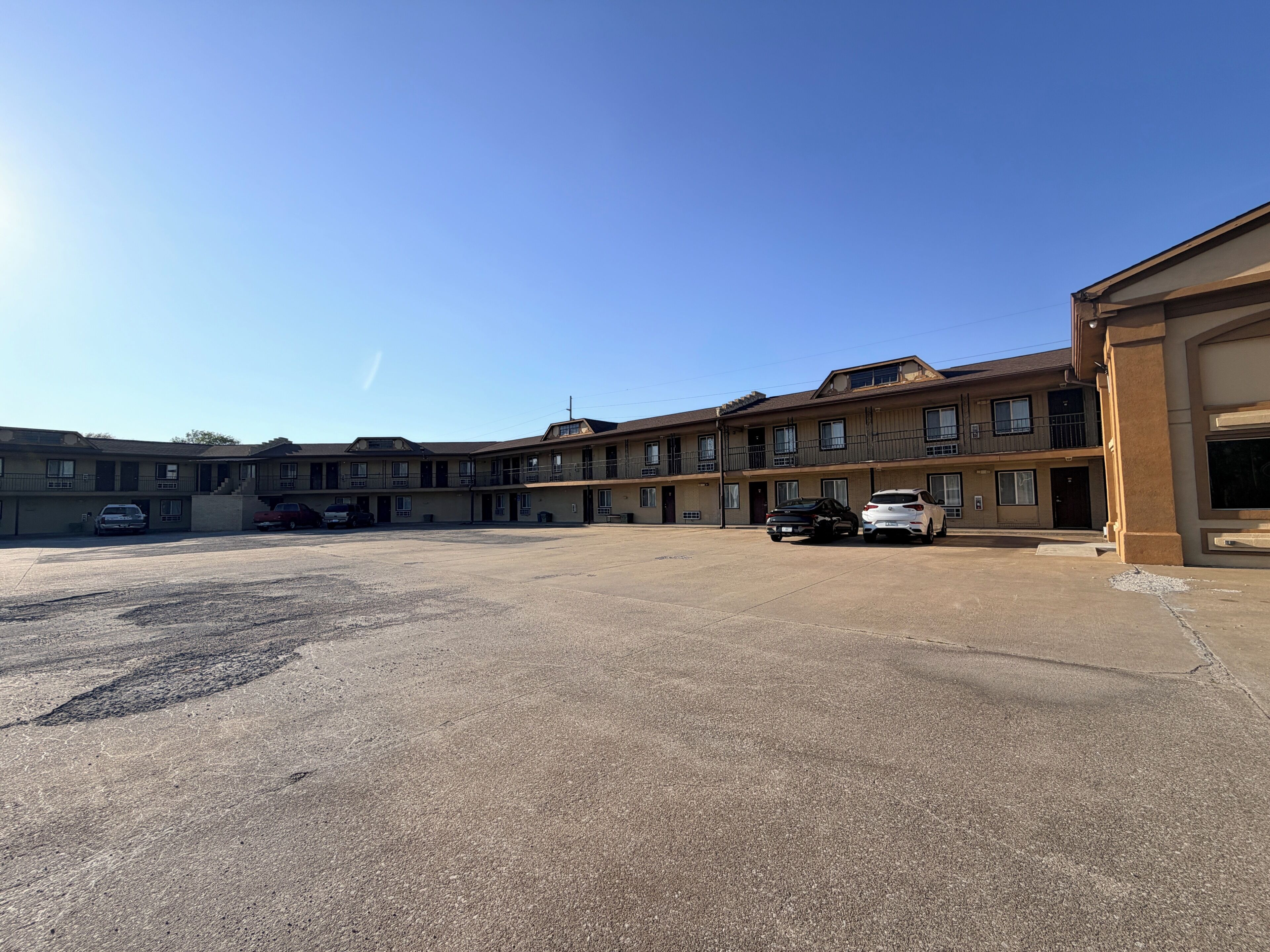 A multi-story motel with a parking lot, several cars parked, and a clear blue sky.