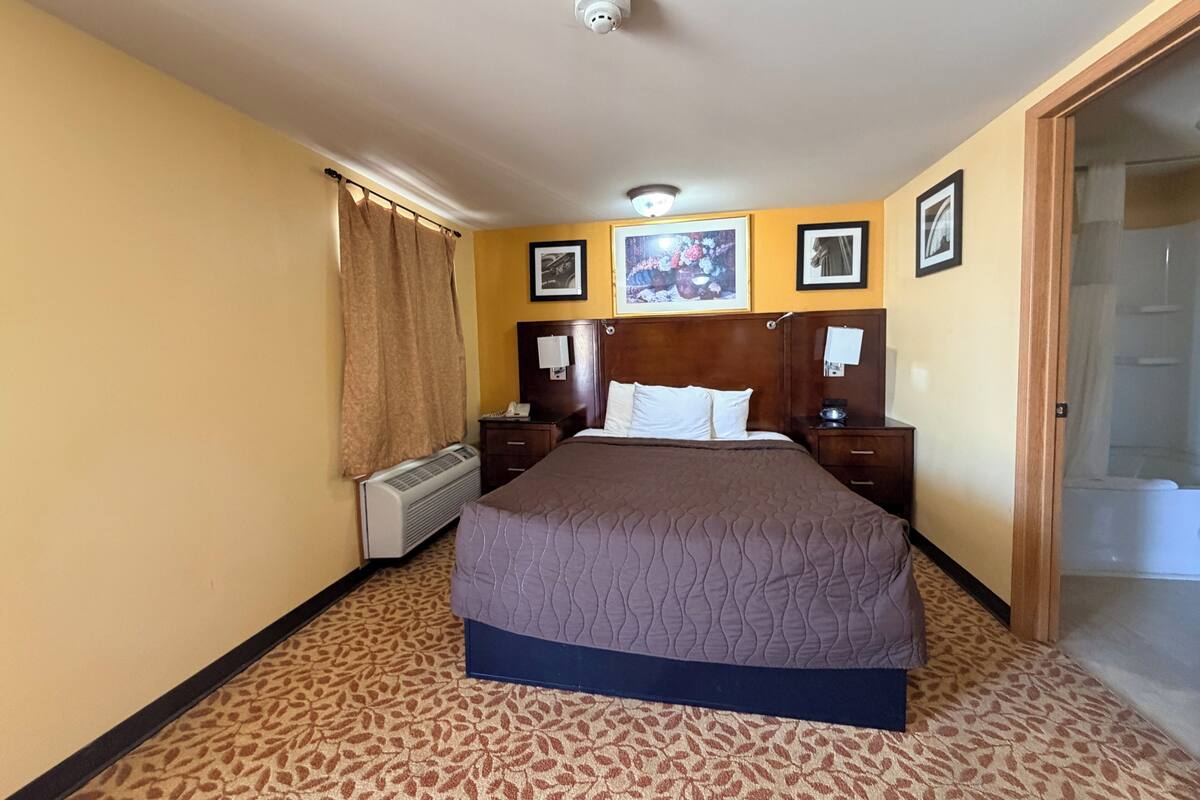 Habitación de hotel con cama, mesita de noche, teléfono, calentador en la pared y puerta que da a un baño.
