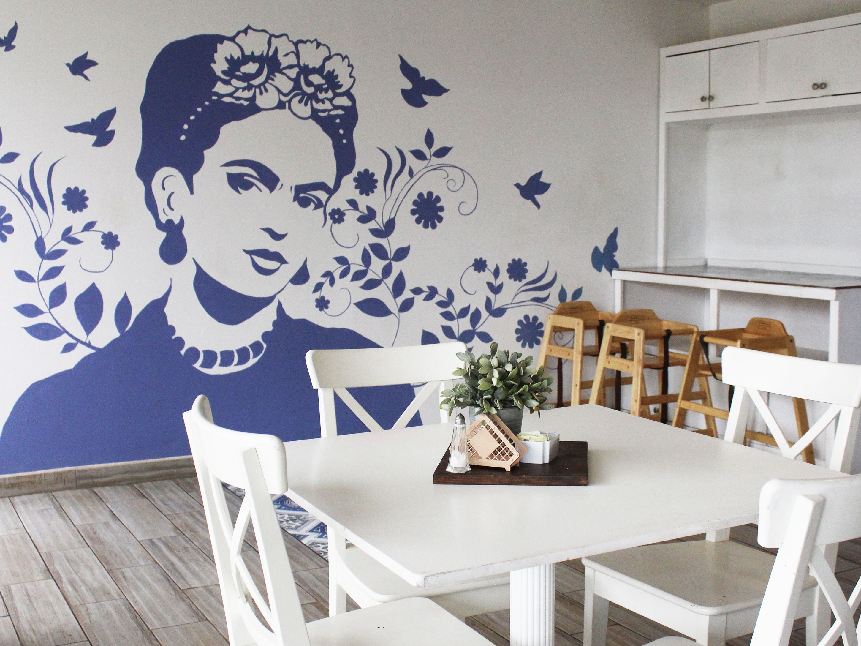 Un área de comedor con una mesa y sillas blancas, un mural decorativo y una planta pequeña sobre la mesa.