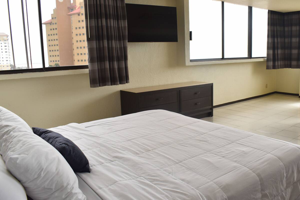 A hotel room with a large bed, a dresser, a flat-screen TV mounted on the wall, and a window with curtains.
