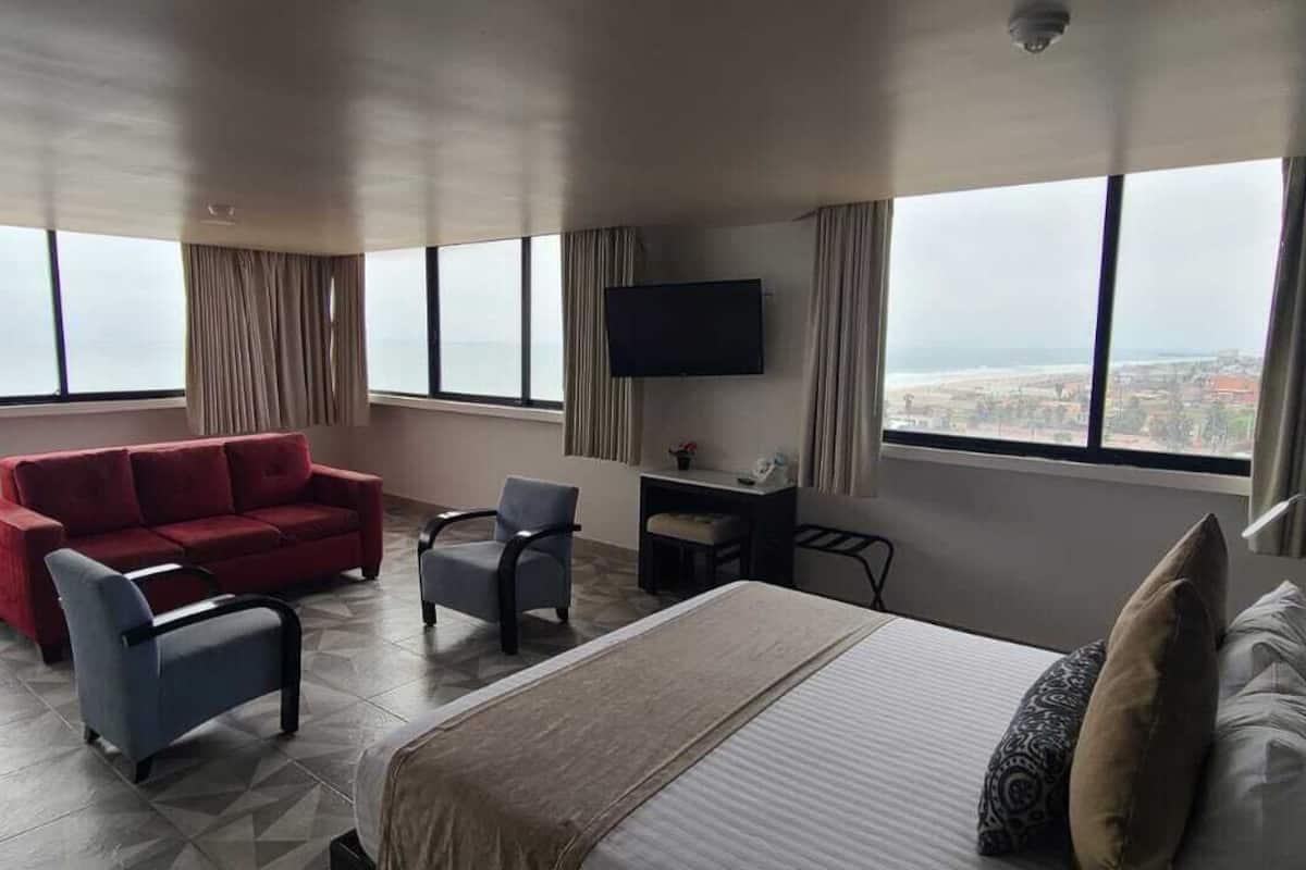 A hotel room with a bed, a red sofa, two armchairs, a desk with a flat-screen TV, and large windows offering a city view.