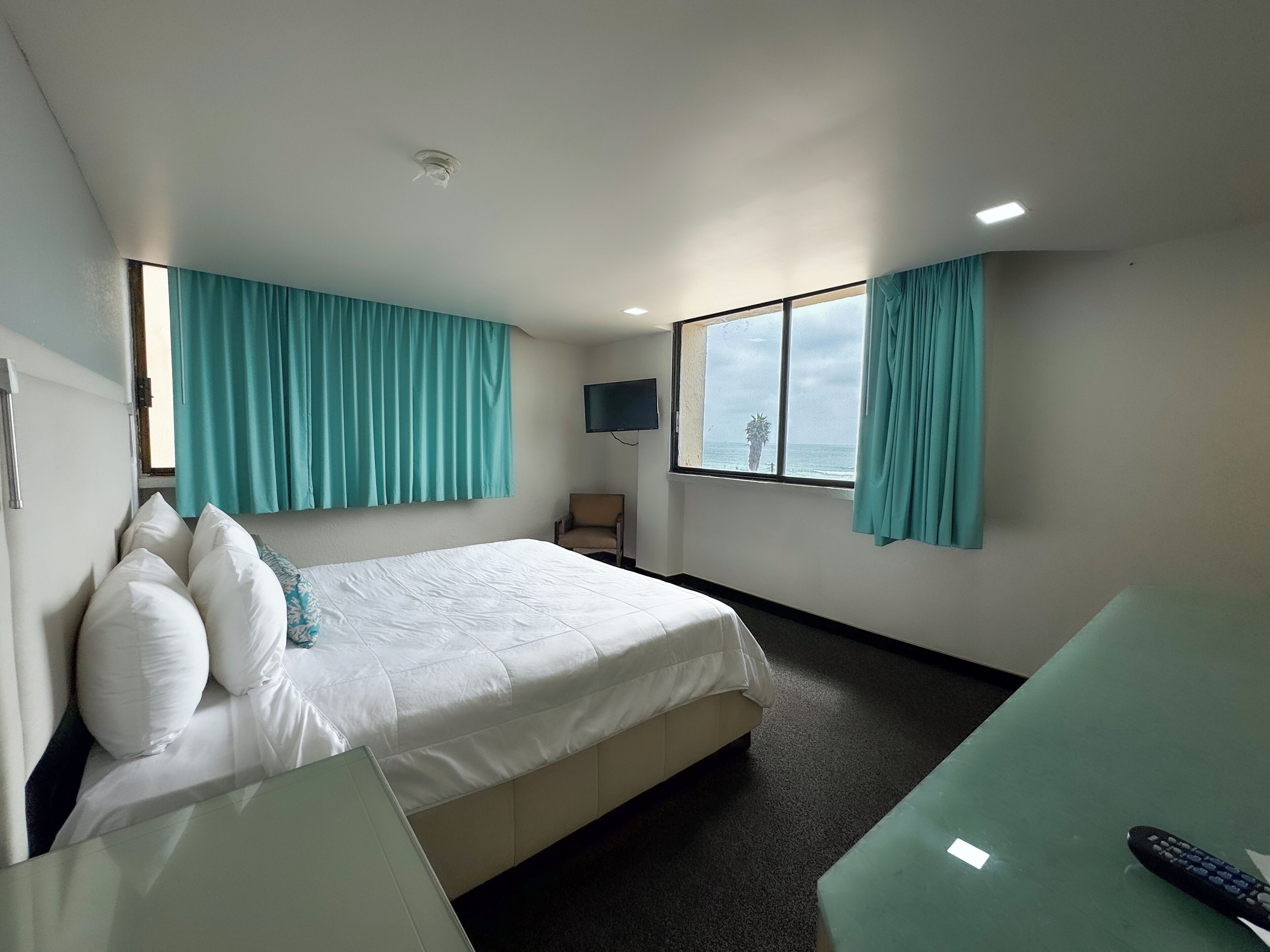 Habitación de hotel con cama, televisor montado en la pared, una silla y ventana con cortinas.