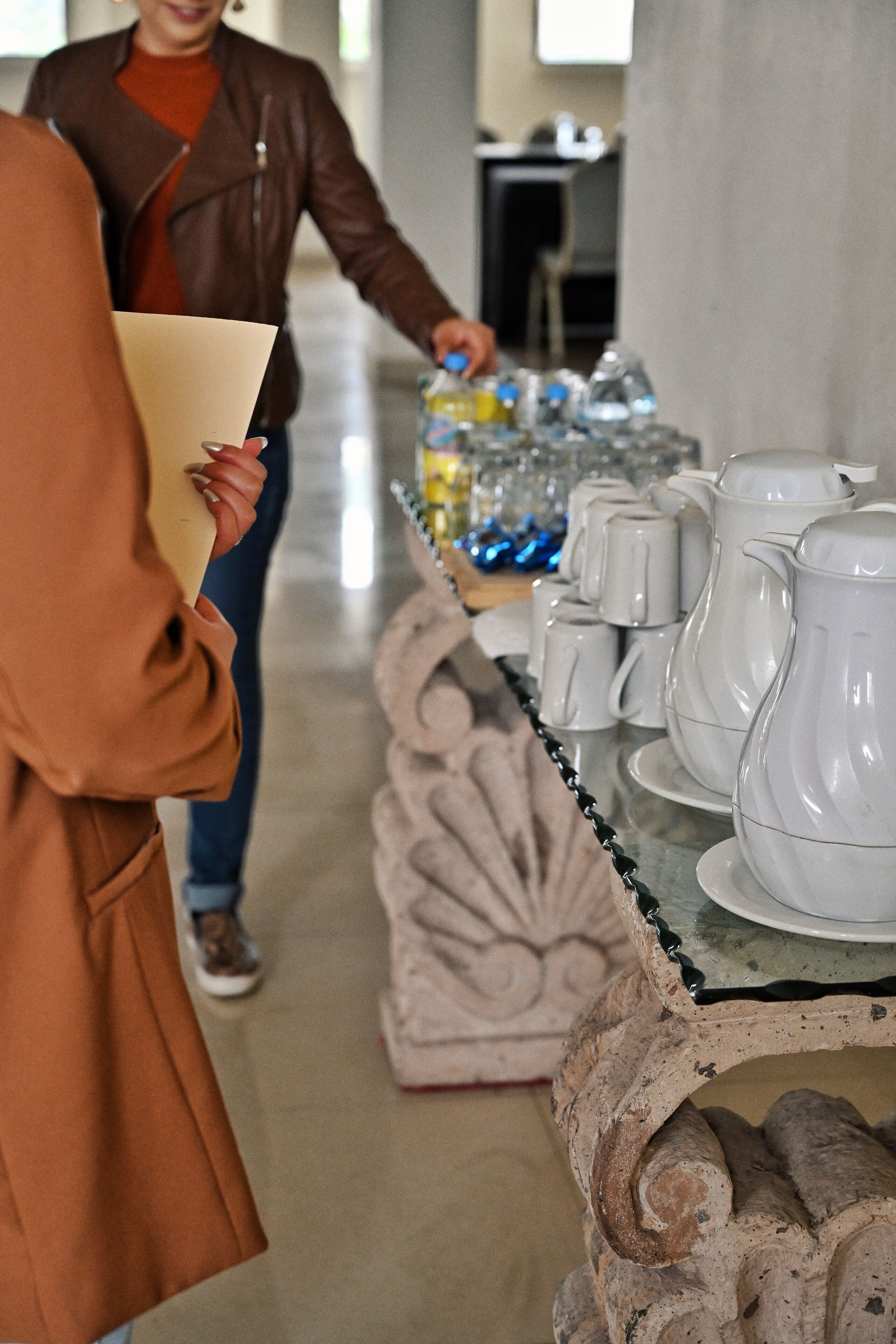 Une personne tenant un dossier dans un couloir, devant une table où sont exposées des tasses et des carafes en céramique blanche.