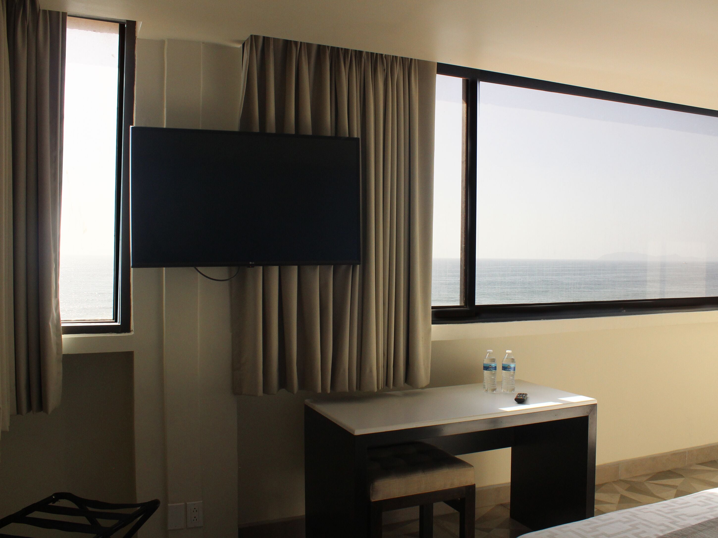 A hotel room with a large window offering a view of the sea, a flat-screen TV mounted on the wall, a small desk with a chair, and a bed.