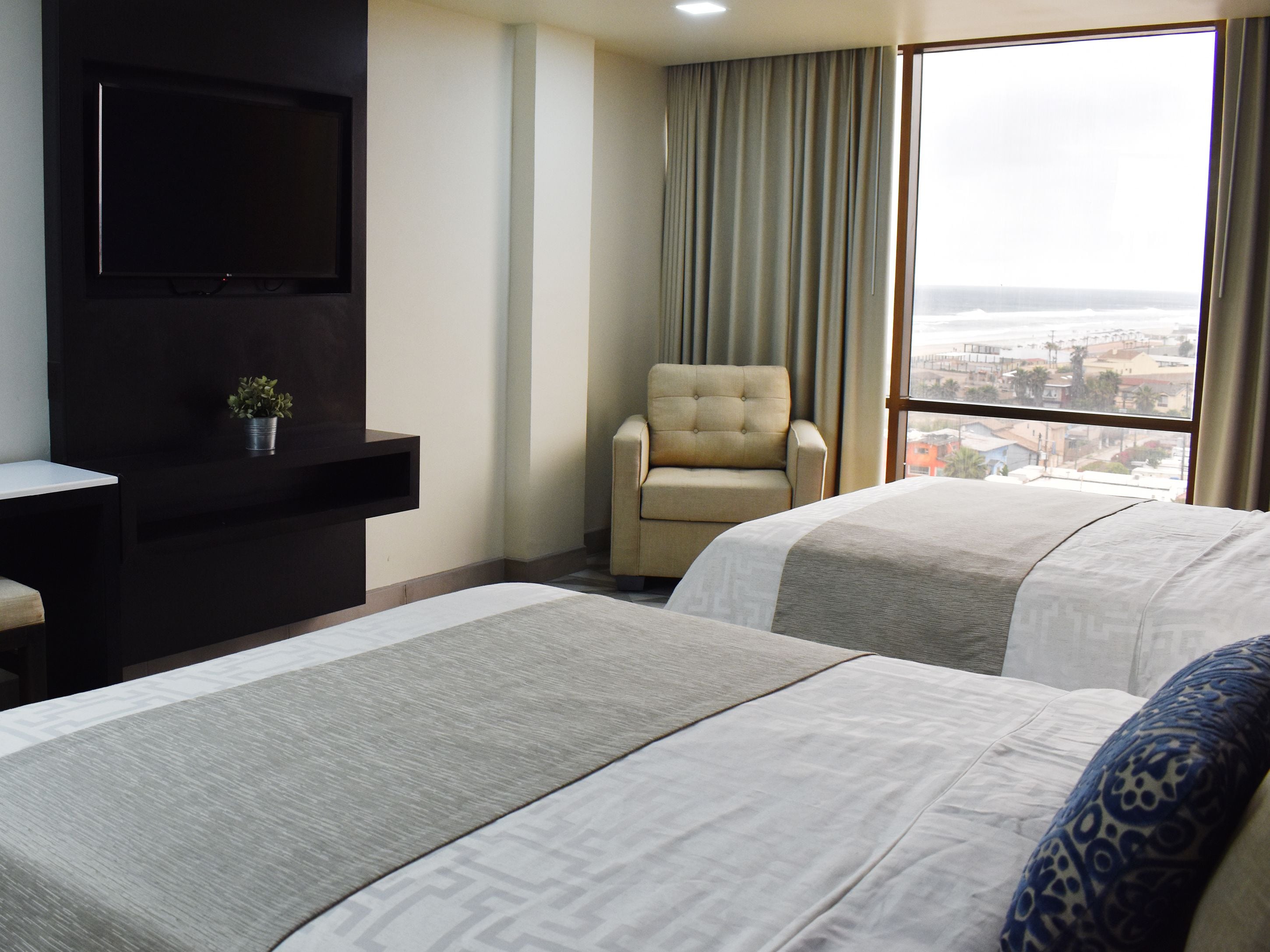 A hotel room with two beds, a TV mounted on the wall, a chair, and a view of the city through the window.