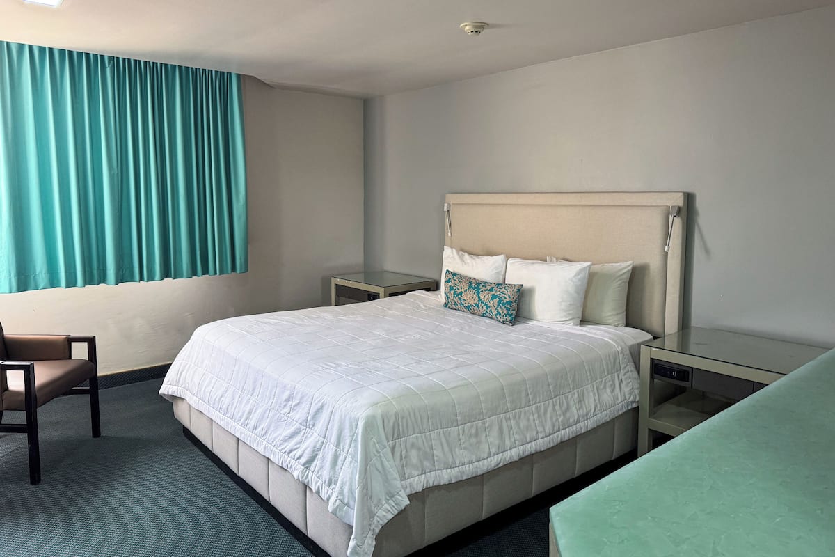A hotel room with a bed, a chair, a nightstand, a bedside table, and a window with curtains.