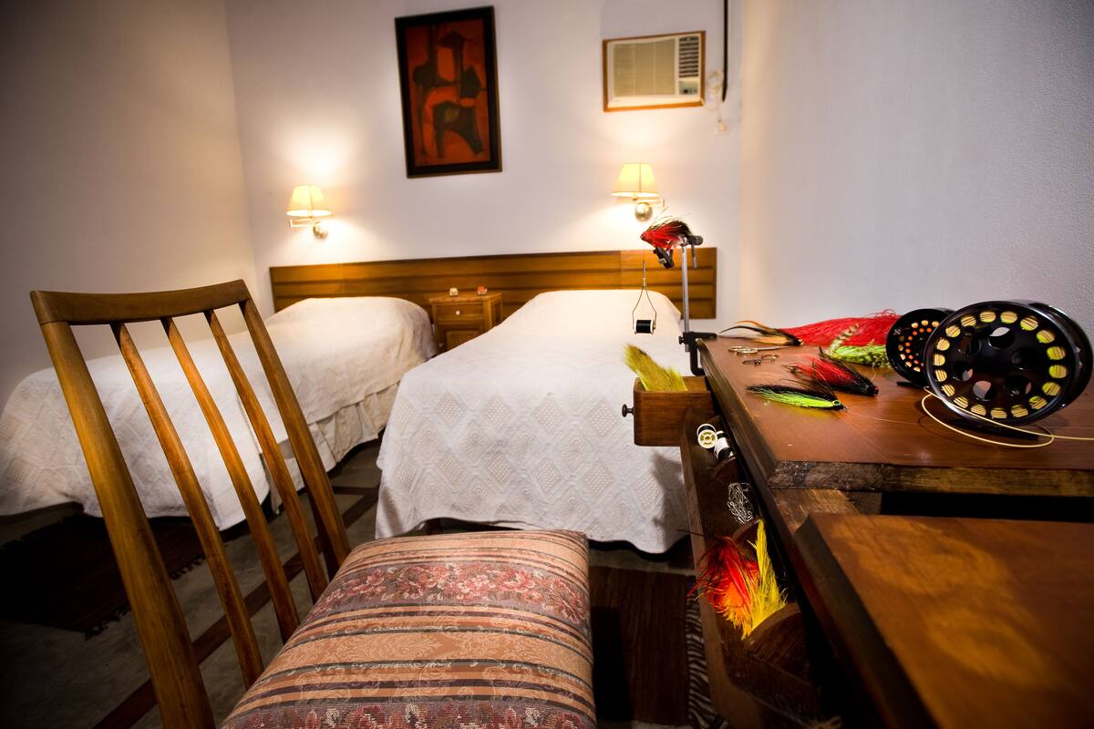 A hotel room with two beds, a wooden chair, a small table with a lamp, and a wall-mounted air conditioner.