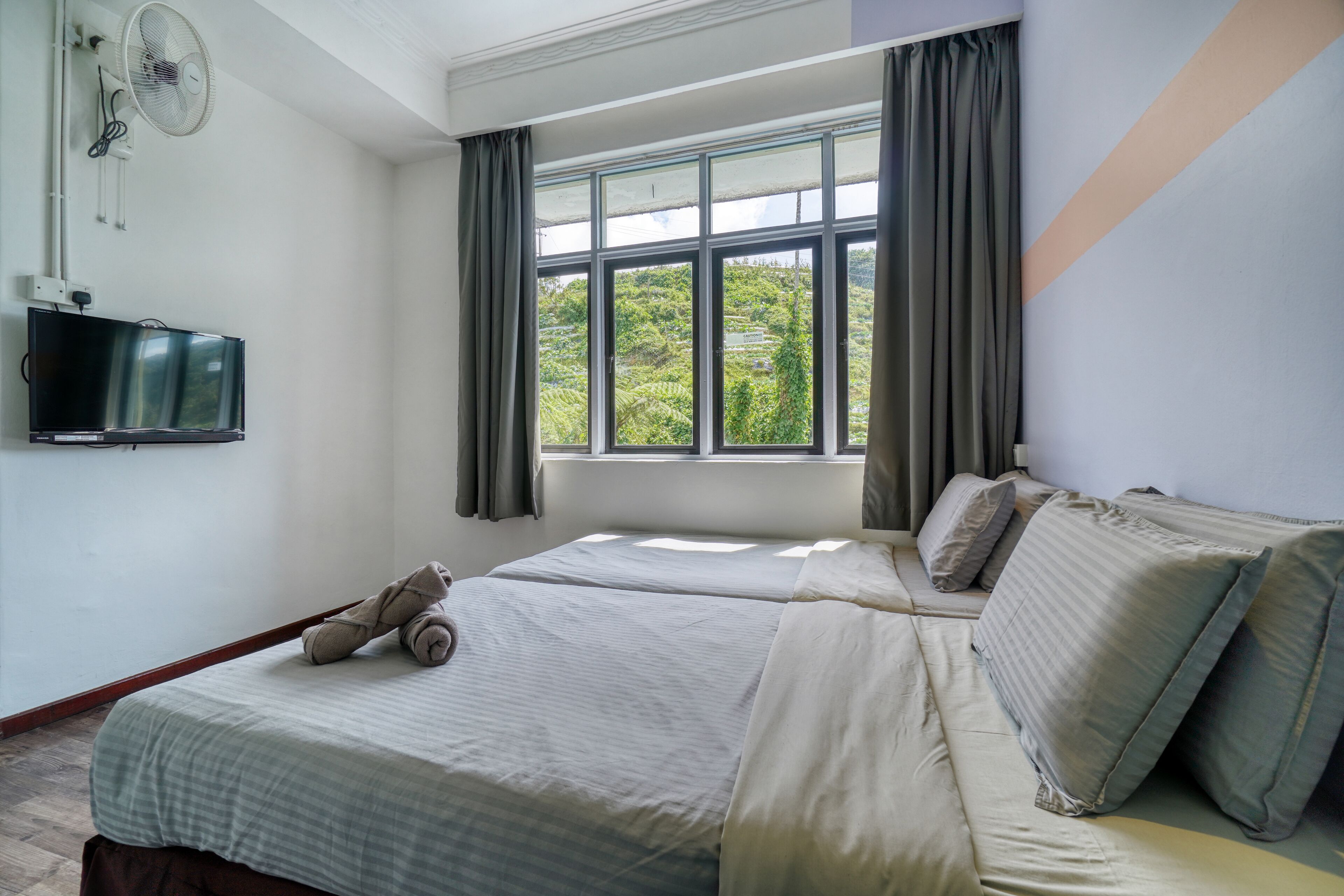 A hotel room with a bed, a television, a window with curtains, and a fan.