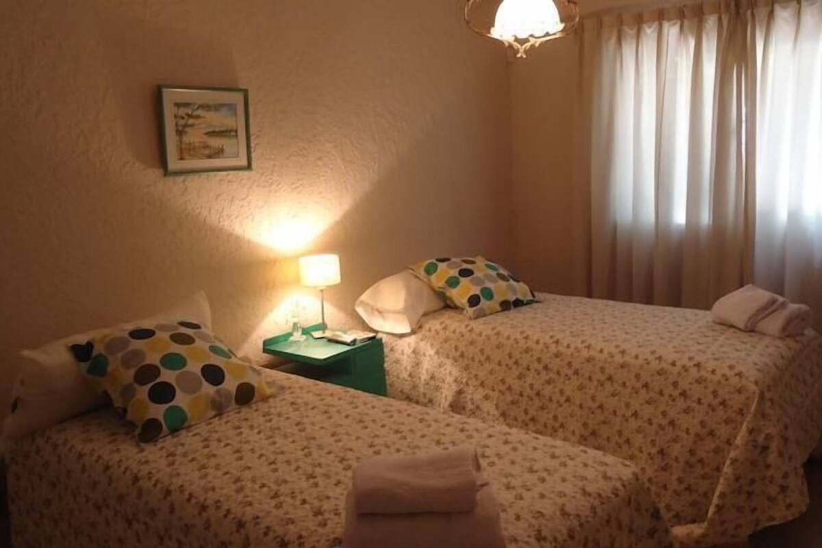 Una habitación de hotel con dos camas, una mesita de noche, una lámpara, un cuadro en la pared y una ventana con cortinas.