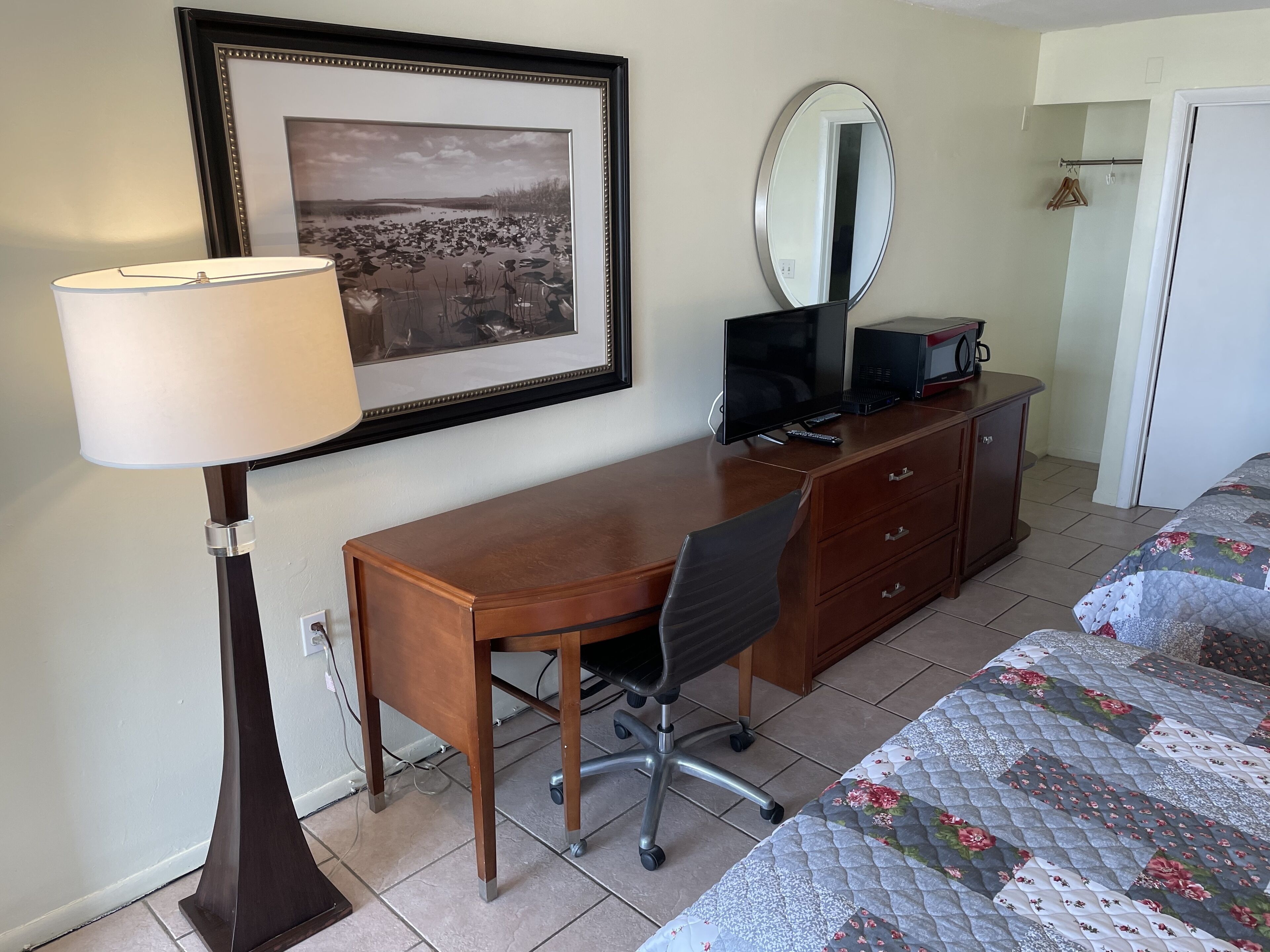 Habitación de hotel con escritorio de madera, una silla, un cuadro enmarco, un espejo y una cama con colcha floral.