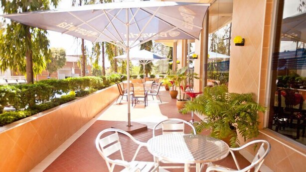 An outdoor patio area with white metal chairs and tables, covered by large umbrellas, and surrounded by potted plants and a well-maintained hedge. - Nyumbani Hotels & Resorts - Moshi (Moshi)