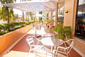 Un patio al aire libre con sillas y mesas de metal blanco, cubierto por grandes sombrillas y rodeado de macetas con plantas y un seto cuidado.