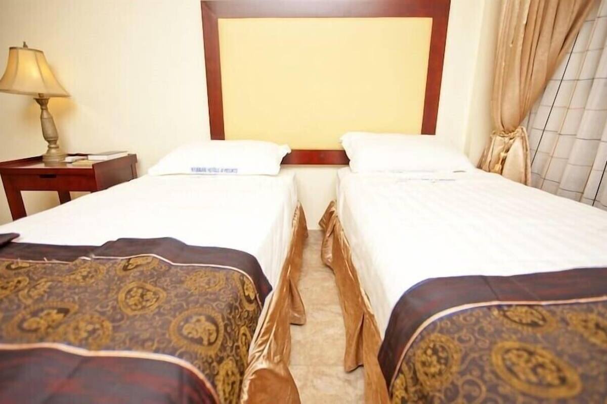 Habitación de hotel con dos camas individuales, una mesita de noche con lámpara y una ventana con cortinas.
