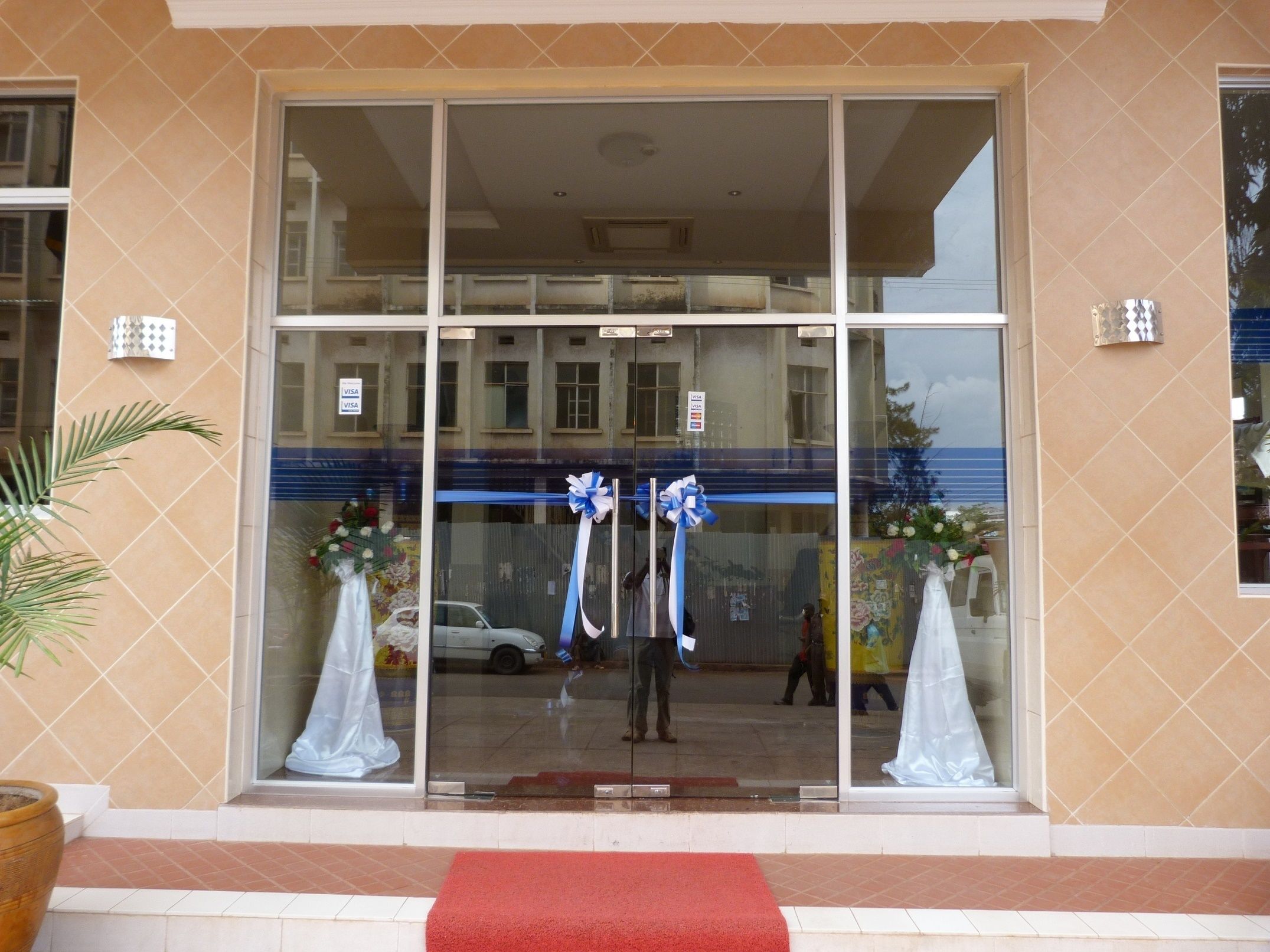 Entrada de un edificio con puertas de cristal grandes, una alfombra roja y dos lazos azules.