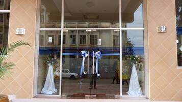 Entrada de un edificio con puertas de cristal grandes, una alfombra roja y dos lazos azules.