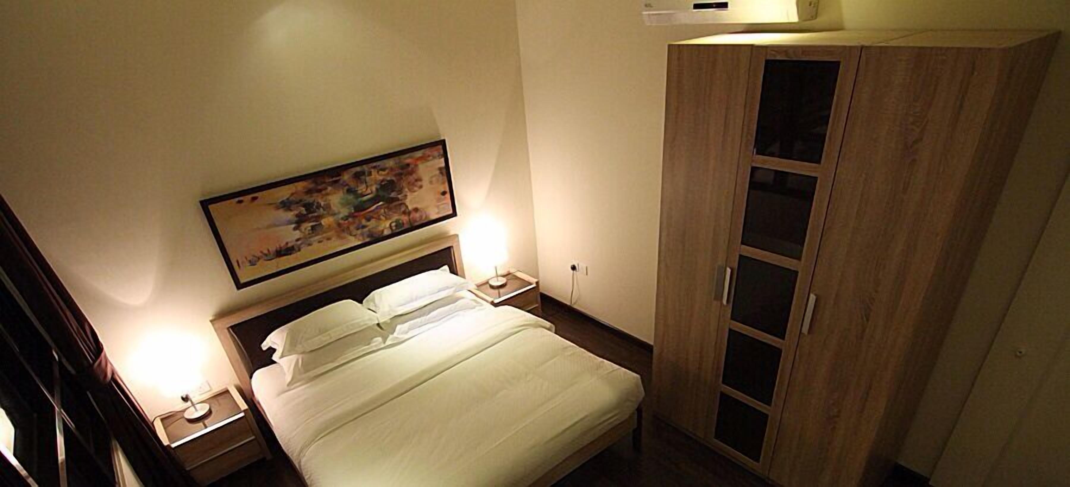 Una habitación de hotel con cama, lámparas de noche, un armario de madera y un cuadro en la pared.