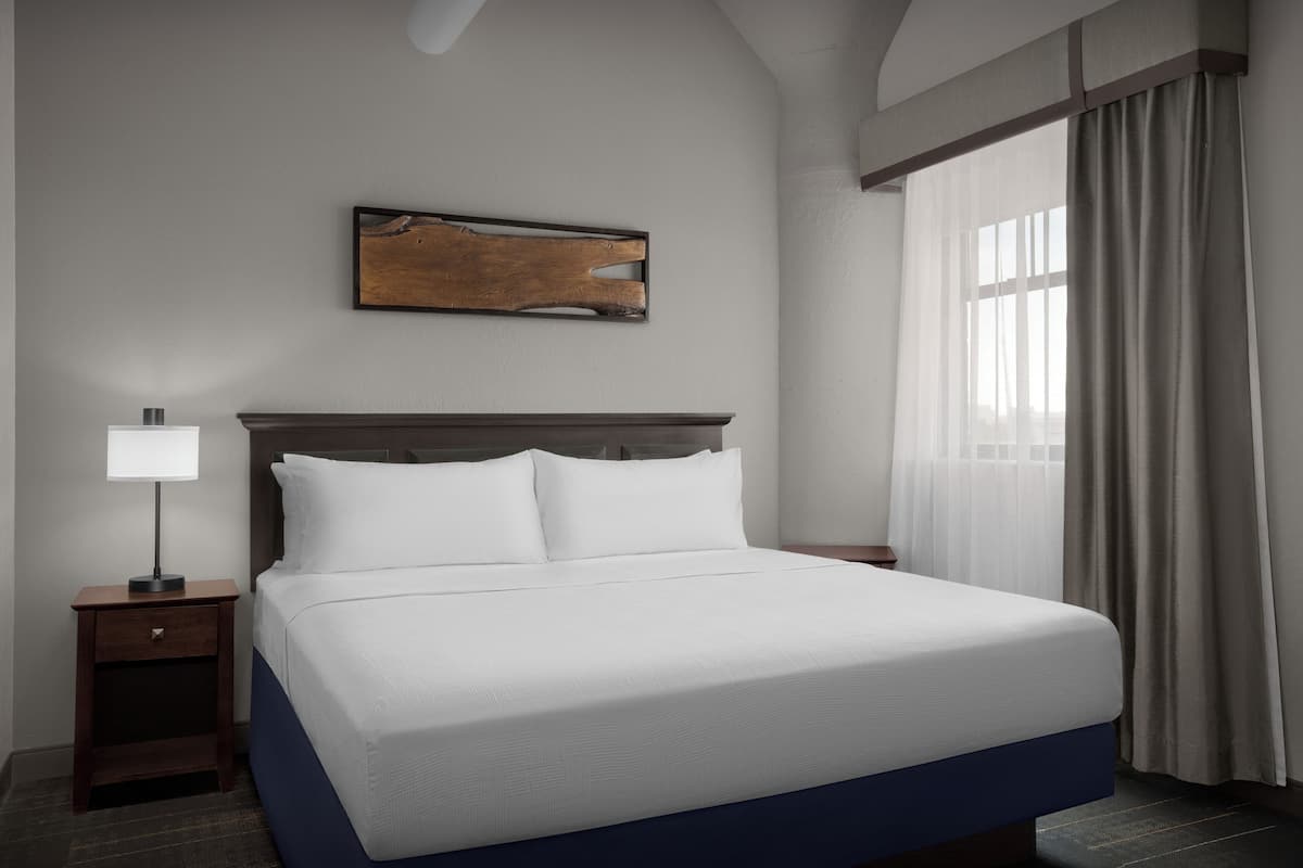 A hotel room with a large bed, a nightstand with a lamp, a wooden headboard, a painting on the wall, and a window with curtains.