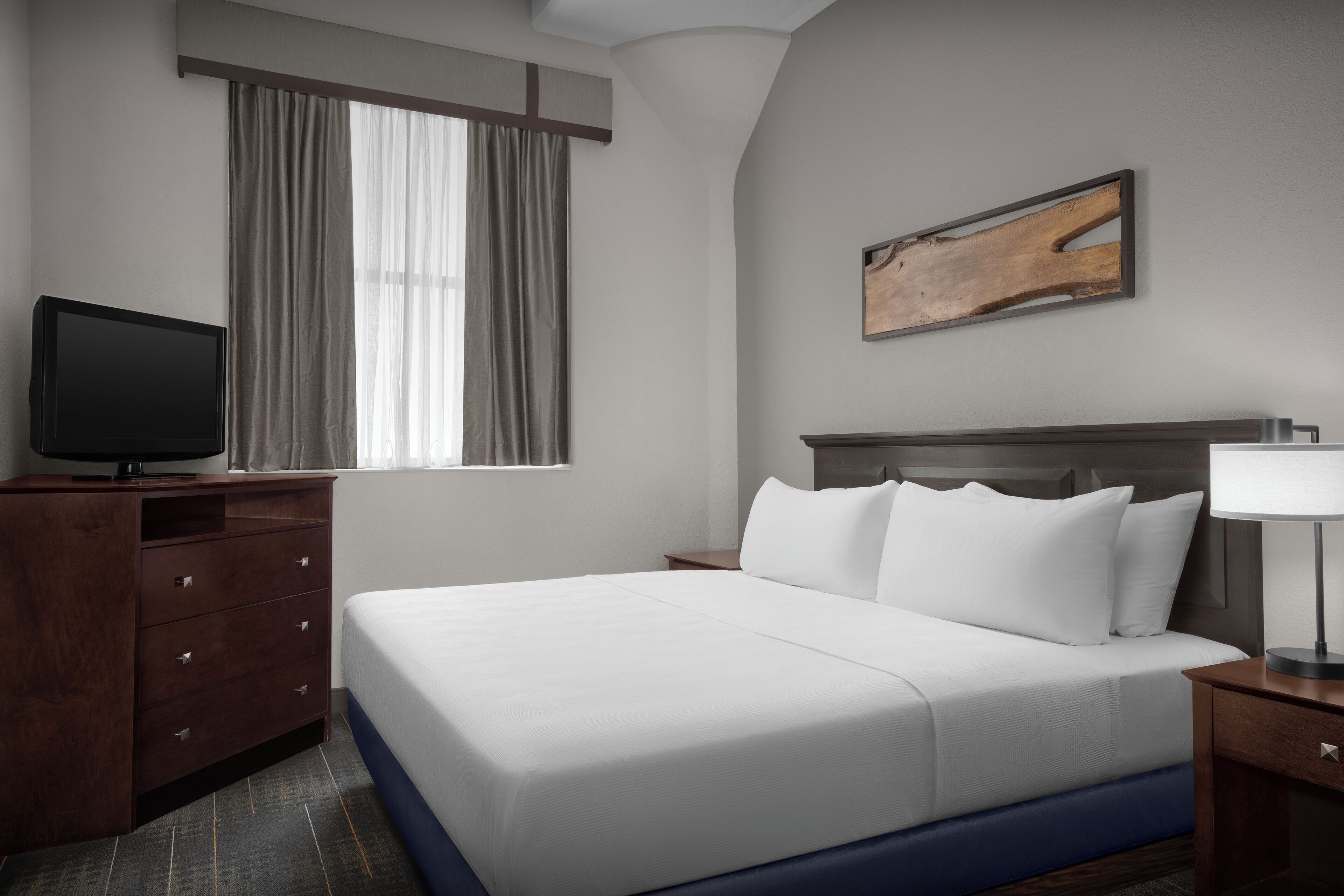A hotel room with a bed, a television, a nightstand, a lamp, and a painting on the wall.
