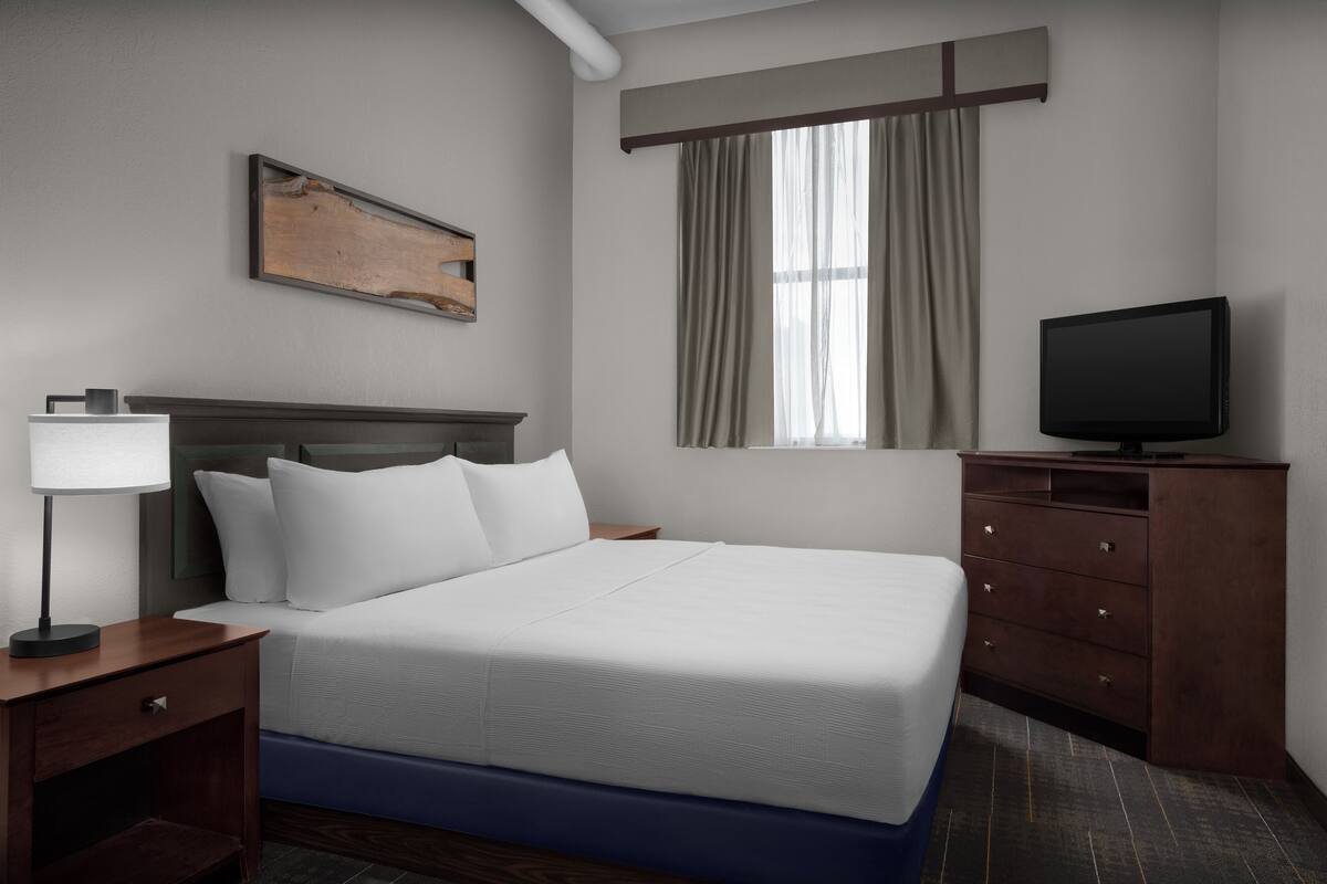 A hotel room with a bed, a TV, a wooden headboard, a nightstand, a lamp, and a window with curtains.