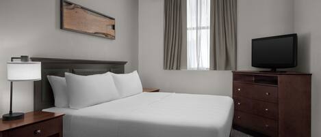 Una habitación de hotel con cama, televisión, cabecera de madera, mesita de noche, lámpara y ventana con cortinas.